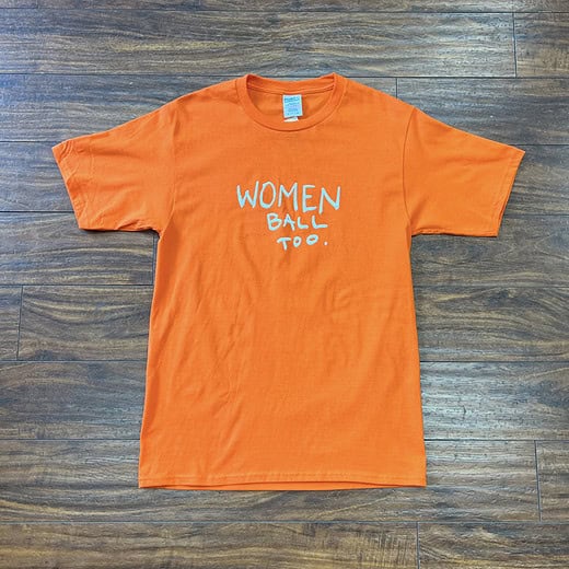Orange "Women Ball Too" t-shirt laid flat on a wooden floor, size small.